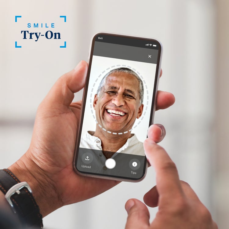 A person using a smartphone to view the Aspen Dental Smile Try-On app, featuring a digital preview of a smiling man. The app interface enables users to explore virtual smile options.