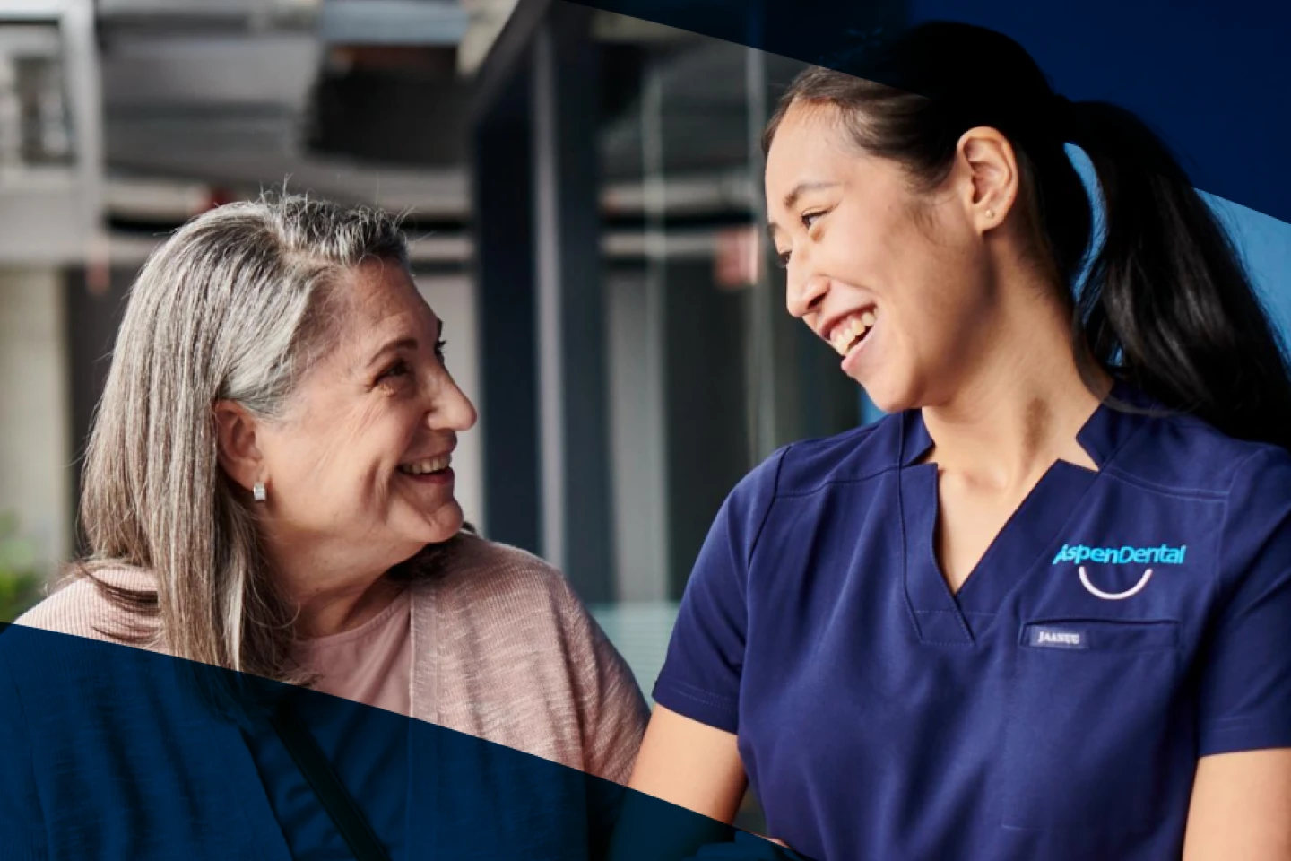 An older woman and a young woman in an Aspen Dental scrubs smile at each other.